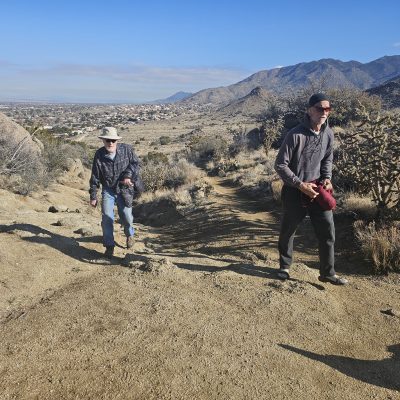 Two hikers on desert trail with mountain views.